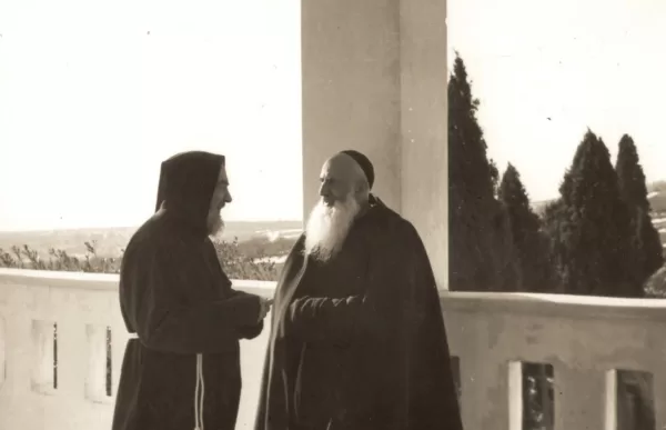 Dwaj zakonnicy rozmawiający na klasztornym krużganku; cień sosnowych drzew; kontekst religijny; obcowanie w ciszy i skupieniu; duchowość katolicka.